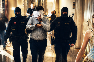 Two armed guards in black gear escort a man who covers his face with a sheet of paper down a hallway, while a glamorous woman in a sparkling dress watches from the right.