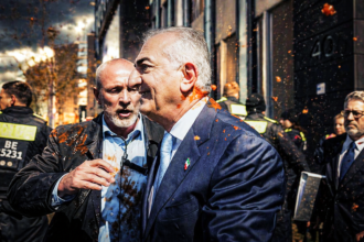 Two men in suits embrace amid orange confetti on a city street, with security in the background.