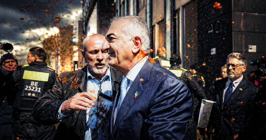 Two men in suits embrace amid orange confetti on a city street, with security in the background.