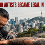 Tattoo artist in a mask and gloves tattoos a client’s forearm; city skyline and a traditional Korean temple rise in the background.