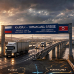 Border bridge at sunset showing Russian and North Korean flags, with a blue sign reading KHASAN – TUMANGANG BRIDGE and trucks crossing over a river.