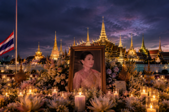 Framed portrait of Thailand's queen amid flowers and candles at night, with the grand palace temple in the background.