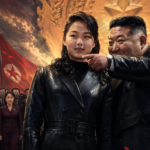 Two people in black leather jackets stand in the foreground as a crowd of officials watches, red flags waving behind them under a dramatic sky.